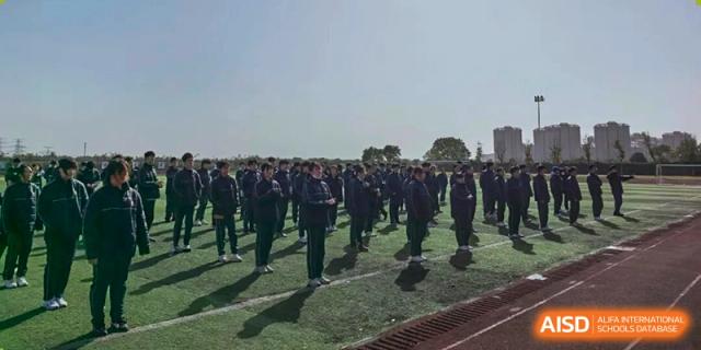 A large crowd of students in dark uniforms is lined up on a sports field's track under a bright sky, possibly for an assembly or drill formation at Maple Leaf International School Shanghai.