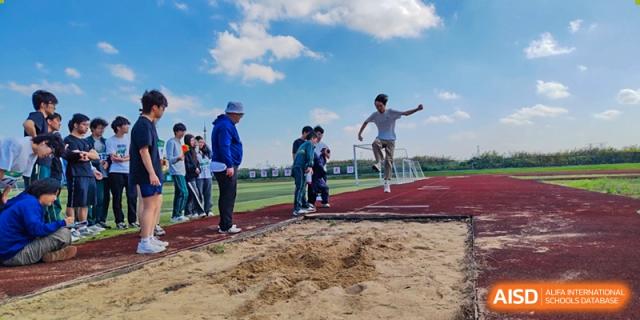 A student is captured mid-air during a long jump attempt into a sand pit on a bright day, observed by other students at Maple Leaf International School Shanghai.