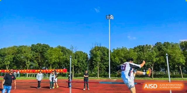 Students practice volleyball on a red sports track next to a high stadium light pole and green trees under a bright blue sky at Maple Leaf International School Shanghai.