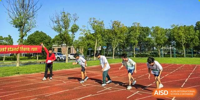 Two students practice the baton exchange for a relay race on a red running track with green trees and campus buildings in the background at Maple Leaf International School Shanghai.