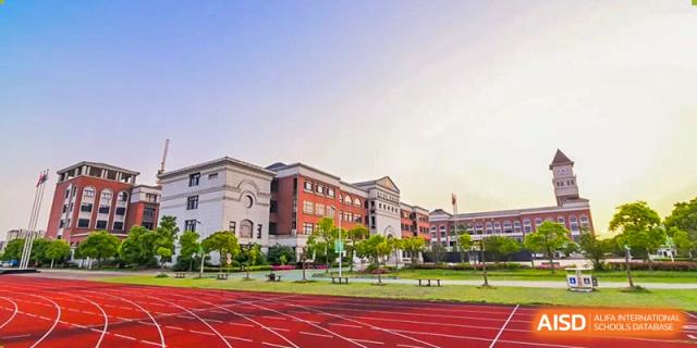 A wide shot of a large brick school building overlooks an empty red running track and a green field under a clear, bright sky at Maple Leaf International School Shanghai.