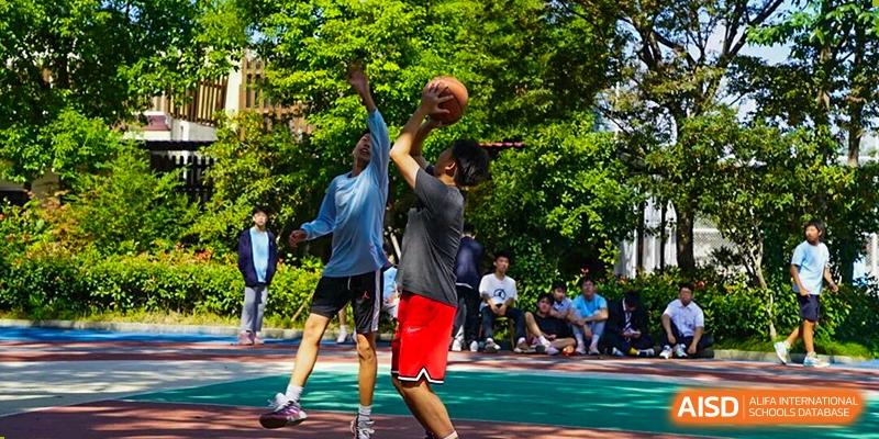Two students playing a competitive game of basketball outdoors on a sunny day, jumping for a shot under the basket, with spectators watching, Living Word Shanghai.