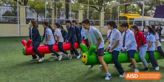 Students in school uniforms participating in a cheerful outdoor team-building activity, riding a large green caterpillar toy on a grassy field, Living Word Shanghai.