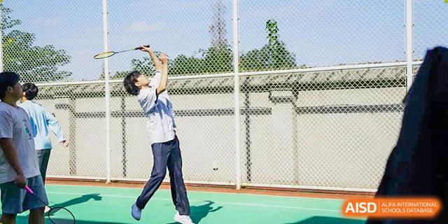 A student in a light-colored uniform practicing an overhead serve during a game of tennis on the school's outdoor court on a bright, sunny day, Living Word Shanghai.