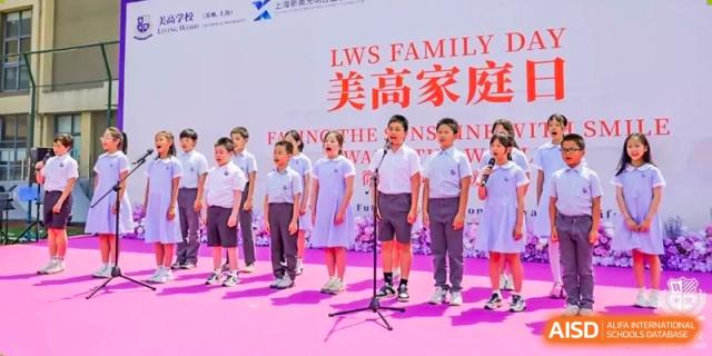 Students in white polo shirts and navy shorts singing on a stage with microphones for the LWS Family Day event banner displayed behind them, Living Word Shanghai.