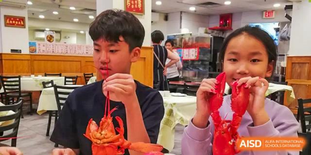 A male student in a red shirt holds up a small cooked lobster with both hands in a busy indoor dining environment. Living Word Shanghai