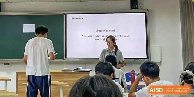 A teacher standing in front of a class, presenting a slide titled "10 min to read" on a projector screen to attentive students in a lecture hall at Jian Ping High School.