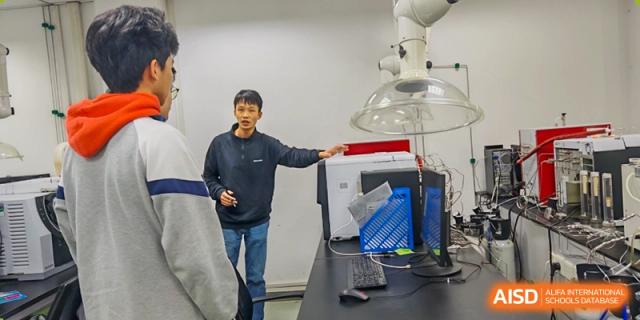 A student presenting or explaining a computer setup and a scientific apparatus in a laboratory to a classmate at Jian Ping High School.