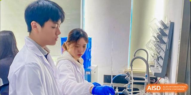 A female student wearing gloves and a lab coat carefully performing a procedure with glassware while being observed by two male students at Jian Ping High School.