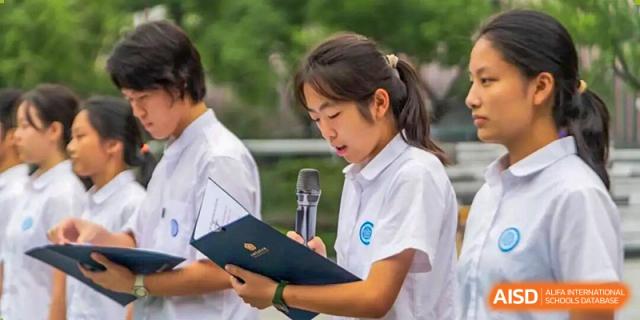 A female student in a white uniform focuses intently while writing notes on a document during an outdoor ceremony or event at Jian Ping High School.