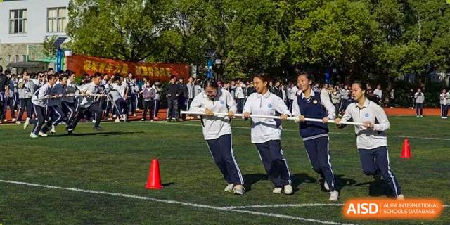 Students in white and blue uniforms strain while participating in a spirited tug-of-war match on a school sports field with spectators nearby, Jian Ping High School.