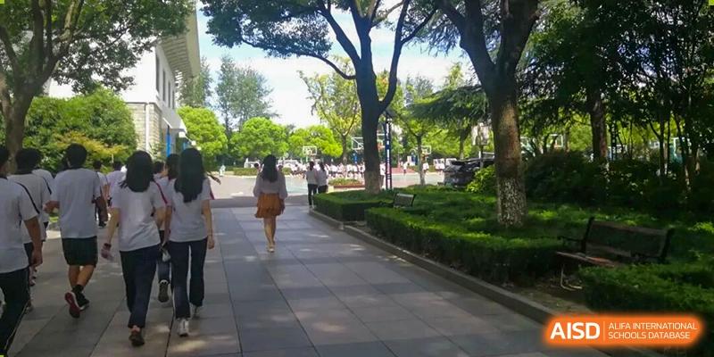 A pathway in a park or campus setting with lush green trees and shrubbery, a person walking away in the distance, Jian Ping High School.