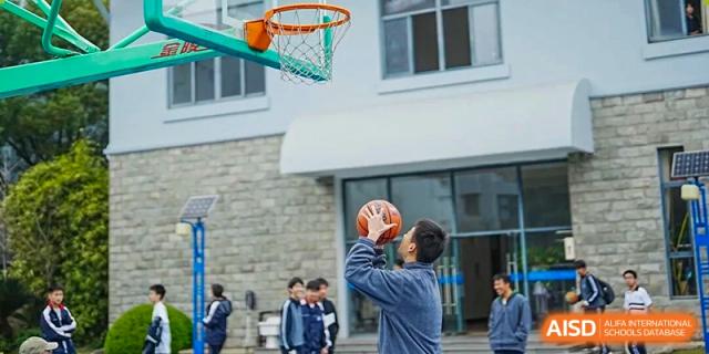 A student attempting a jump shot with a basketball on an outdoor court, with other students and a building in the background, Jian Ping High School.