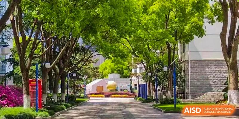 A wide, paved path on a university or school campus lined with tall, lush green trees and manicured lawns, Jian Ping High School.