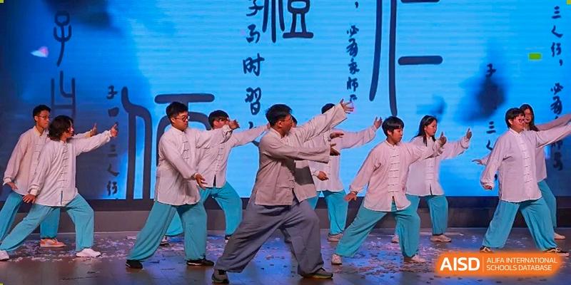 Four male students performing a traditional or interpretive dance in loose, light-colored robes on a stage with a blue screen, Jian Ping High School.