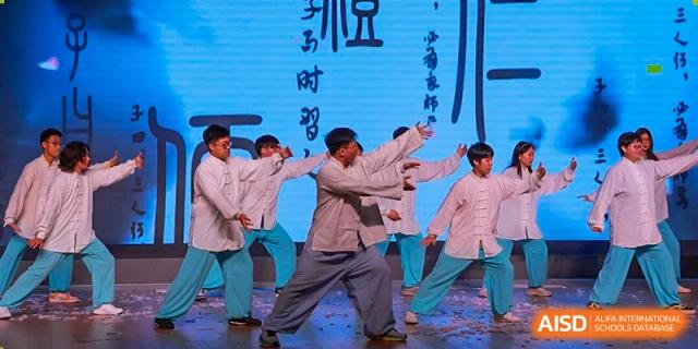Four male students performing a traditional or interpretive dance in loose, light-colored robes on a stage with a blue screen, Jian Ping High School.