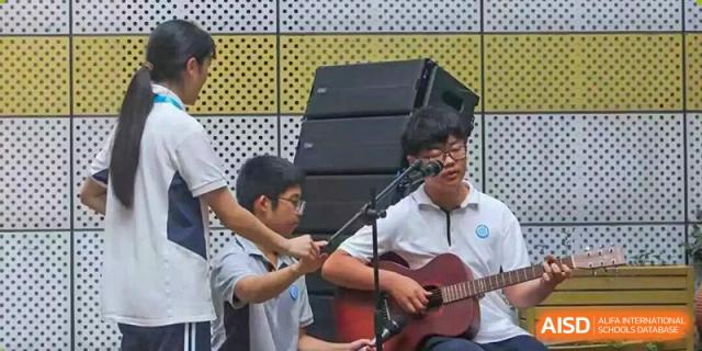 Two male students playing acoustic and electric guitars during an outdoor performance, with large audio speakers visible behind them, Jian Ping High School.