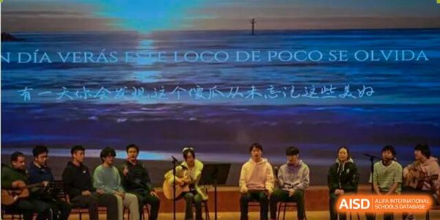 Four students sitting and playing acoustic guitars on a stage in front of a large screen displaying Spanish text, "Este loco de poco," Jian Ping High School.