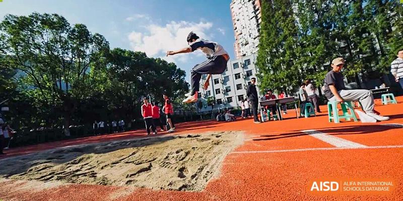 A male student performing a long jump, suspended in the air over the sand pit with an athletic track in the background, Jian Ping High School.