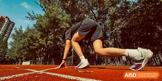 A student crouched low at the starting line of an orange running track, prepared to begin a sprint or race, viewed from a low angle, Jian Ping High School.