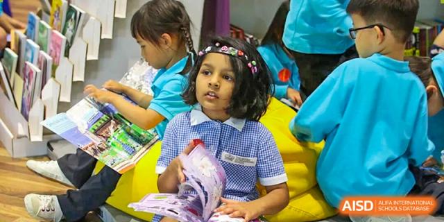 A smiling young girl in a school uniform is sitting down and focused on reading a colorful book in a classroom setting at The British International School Shanghai, Puxi.