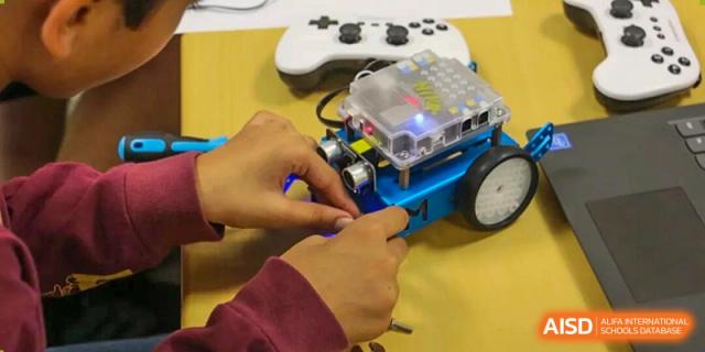 Student hands are working on a small, blue, wheeled robot with electronics during a practical STEM or robotics lesson at The British International School Shanghai, Puxi.