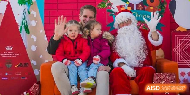 Two young, smiling children sitting comfortably on the lap of Santa Claus during a festive holiday event at the school at The British International School Shanghai, Puxi.