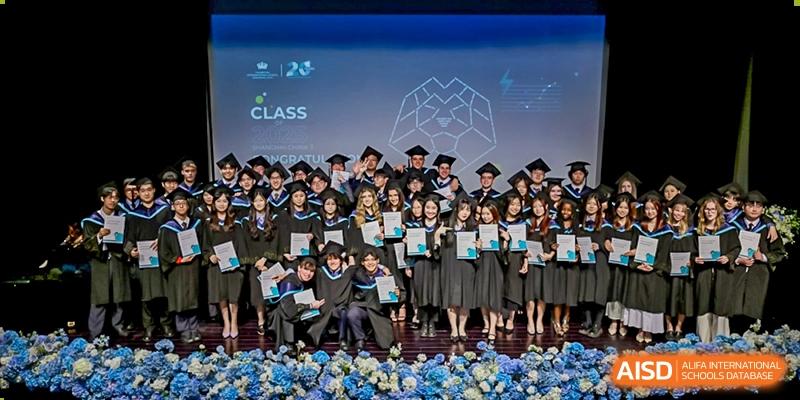 A large group of students in black graduation gowns and caps proudly posing with their diplomas on a stage after a ceremony at The British International School Shanghai, Puxi.
