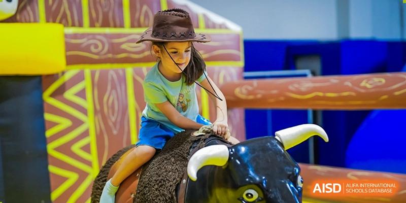 A young girl wearing a cowboy hat laughing while riding a mechanical bull at a fair or festival event at The British International School Shanghai, Puxi.
