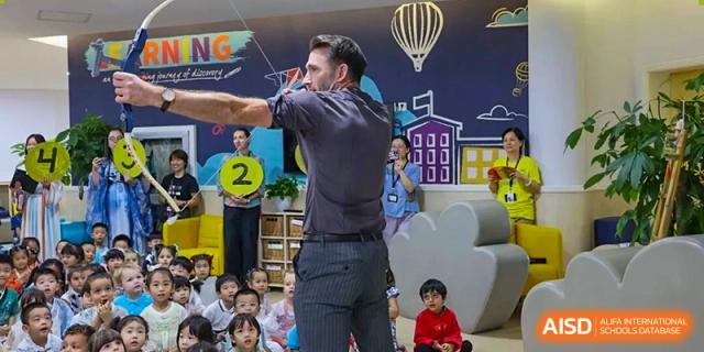 A male teacher demonstrating a theatrical move or pose to an audience of young, seated students during a performance or class at The British International School Shanghai, Puxi.