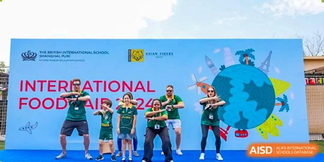 Several individuals in matching green and black uniforms striking a pose on a stage beneath a banner for an 'International Fair' at The British International School Shanghai, Puxi.