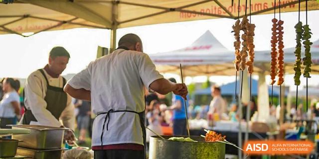 Chef in a white coat and black apron stirring food in a large pot under a tent at an outdoor market event, The British International School Shanghai, Puxi.