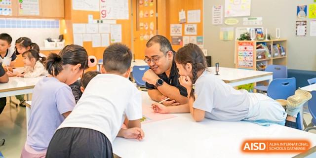 An adult teacher leans over a table helping two young students work on a creative task involving paper and markers in a brightly lit room, Avenues: The World School.