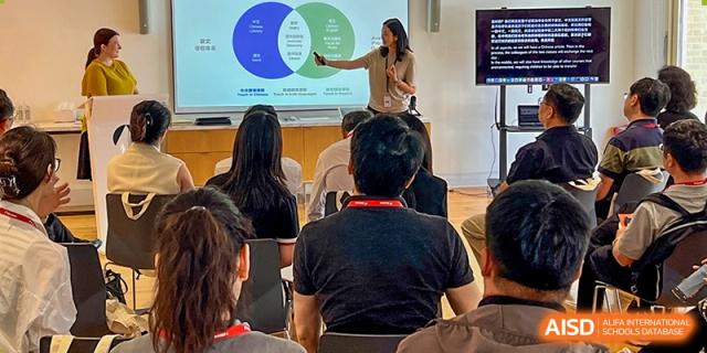 A teacher points to a diagram on a large screen while presenting information to a group of seated students and adults in a classroom, Avenues: The World School.  Middle Row (Left to Right)