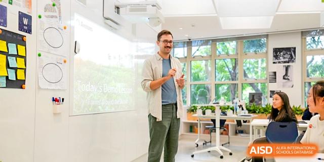 A male teacher stands in front of a projector screen, actively teaching a lesson to his students in a modern, plant-filled classroom, Avenues: The World School.