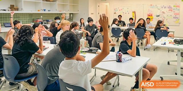 A student raises their hand enthusiastically while seated at a table with other students in a collaborative, well-lit classroom environment, Avenues: The World School.