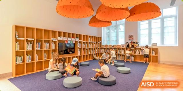 Young students sit on circular blue mats on the floor in a colorful, well-lit library or reading nook with wooden shelves, Avenues: The World School.