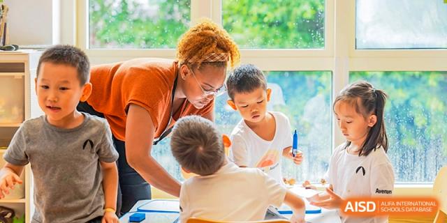 A female teacher with curly hair kneels down, helping two young students with a lesson near a window in a bright classroom, Avenues: The World School.