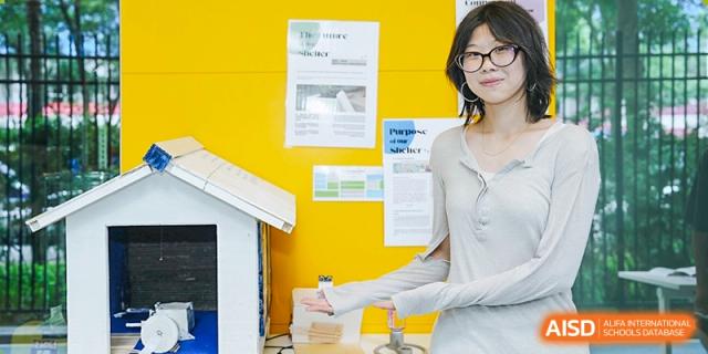 A teacher stands in a classroom, smiling as she gestures toward a whiteboard covered with yellow papers and notes, Avenues: The World School.