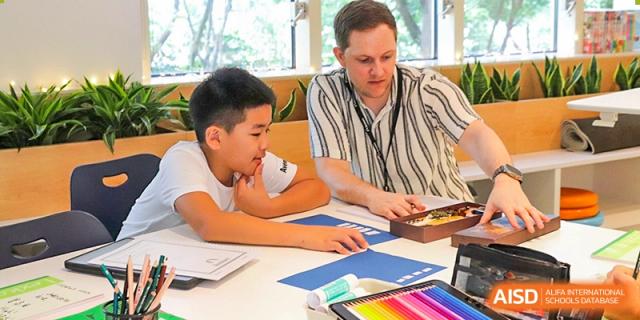 A male teacher sits at a table, actively assisting two students with their school work or projects in a sunny classroom, Avenues: The World School.