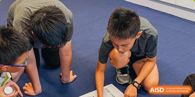 A student wearing dark clothes kneels on a blue mat, intently examining something on the floor, possibly during a practical lesson, Avenues: The World School.