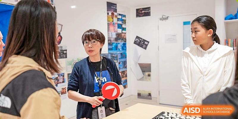 A teacher or staff member wearing glasses and an ID lanyard stands in a classroom or office holding a red sign, Avenues: The World School.