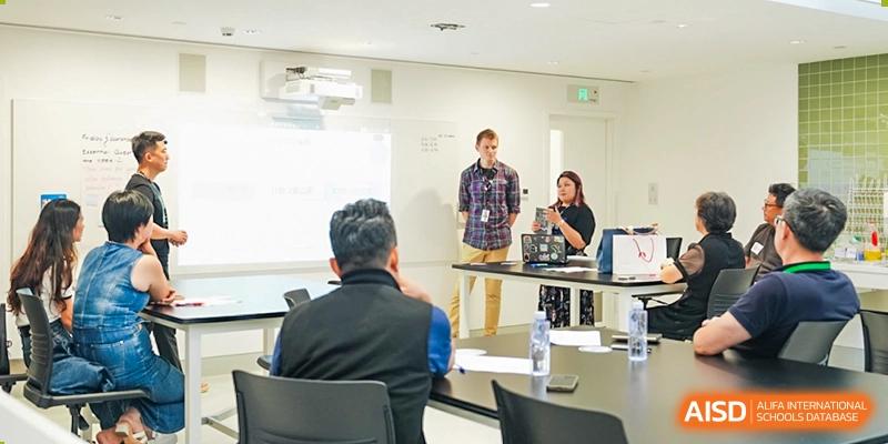 A teacher and staff member stand presenting information to seated adults in a classroom or small conference room, Avenues: The World School.