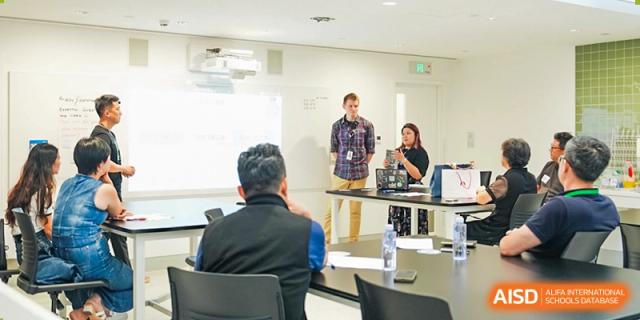A teacher and staff member stand presenting information to seated adults in a classroom or small conference room, Avenues: The World School.