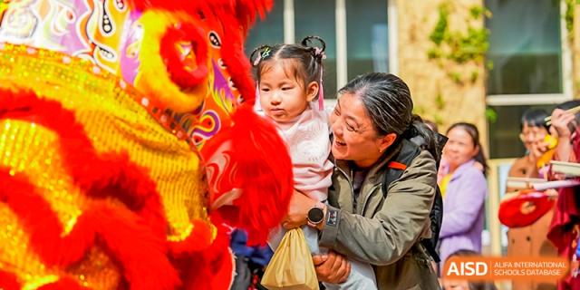 A mother smiles while holding her young child, who is interacting with a colorful lion dance costume or puppet, Avenues: The World School.
