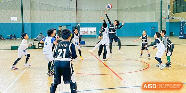 Students in black and white uniforms jump at the net to hit or block a volleyball during an intense indoor game, Avenues: The World School.