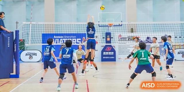 Students in blue uniforms compete in an intense indoor volleyball match, with one player jumping up for a block or spike, Avenues: The World School.
