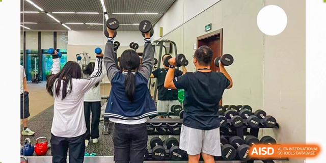 Two students lift dumbbells overhead in a mirrored fitness room, focusing on strength training or physical education, Avenues: The World School.