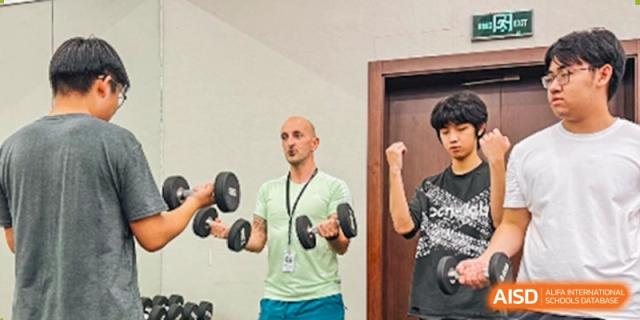 A male instructor demonstrates a dumbbell exercise to a student in a fitness room, while another student observes, Avenues: The World School.