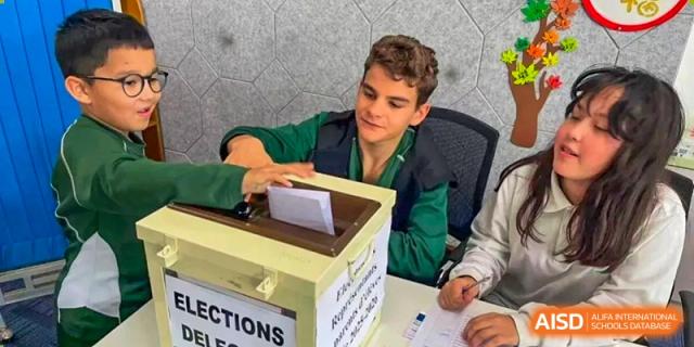 An older student in a green uniform casts a paper ballot into a white voting box labeled "Elections" at Ecole Française Internationale de Wuhan.